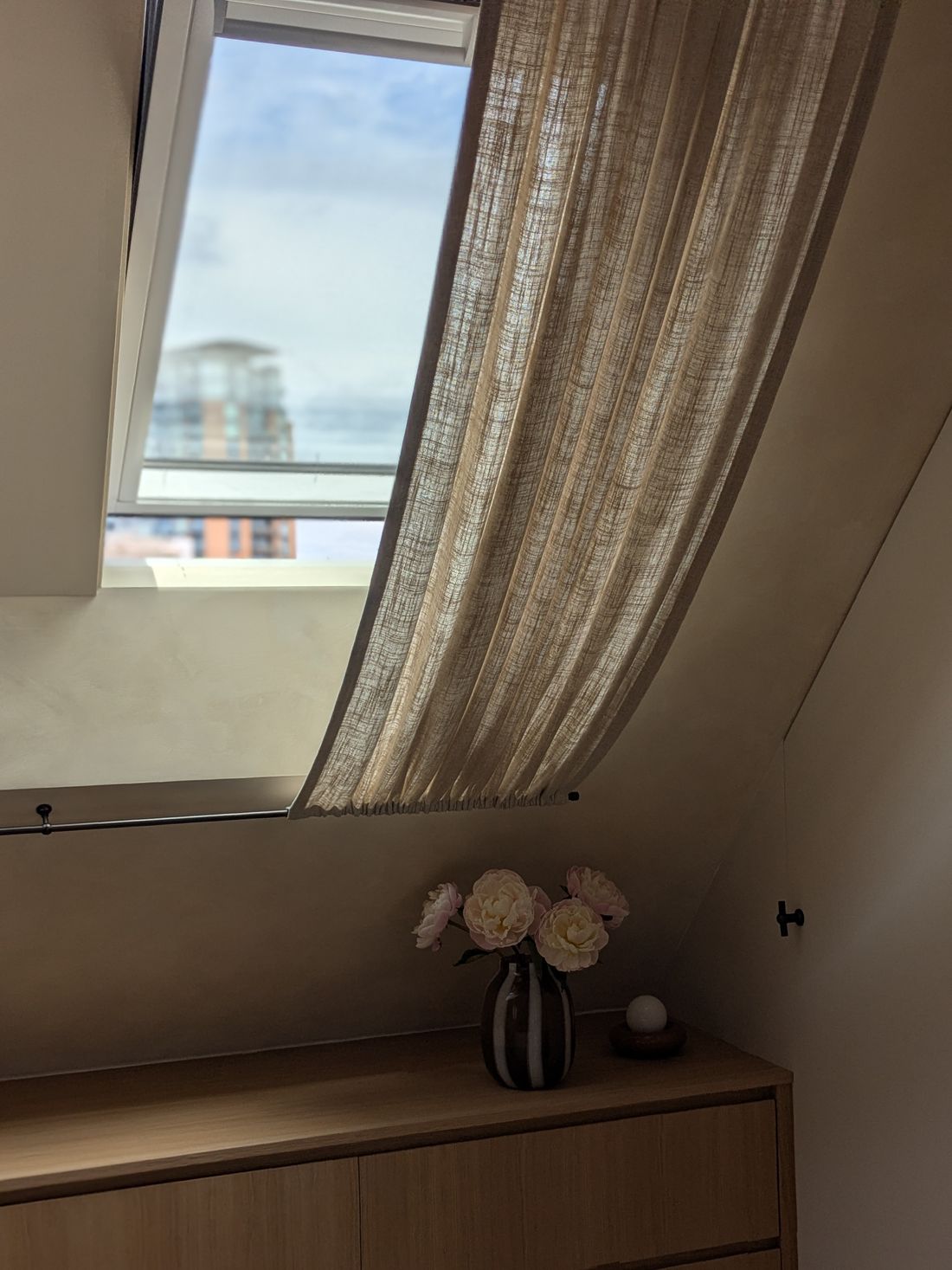 Loft bedroom with a linen curtain draped under a skylight and peonies on an oak chest of drawers, designed by Make My House Home