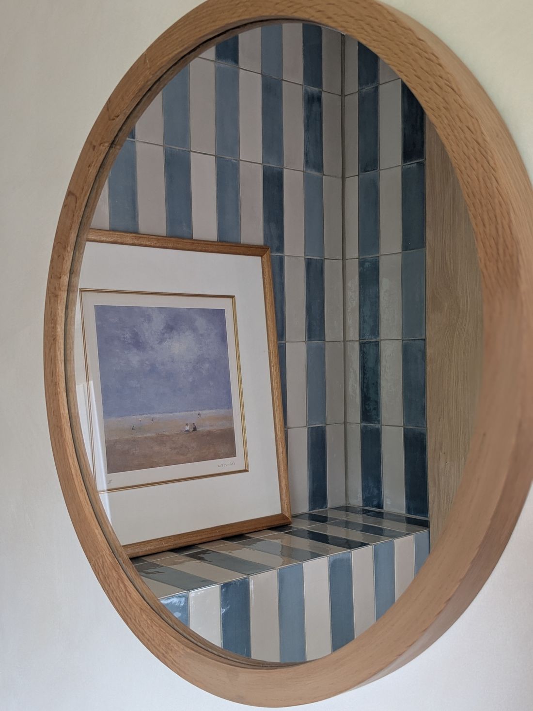 Round oak mirror reflecting a blue and white striped tiled niche with a framed beach print, designed by Make My House Home
